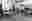Giya Makondo-Wills - Happy Days Black and white photo of a toddler walking across the room of a nursery. The child is wearing a coat and is blurred in the picture due to the movement. There are tables and chairs set up in small groups around the room. There are drawers and shelves around the perimeter of the room and displays on the wall.